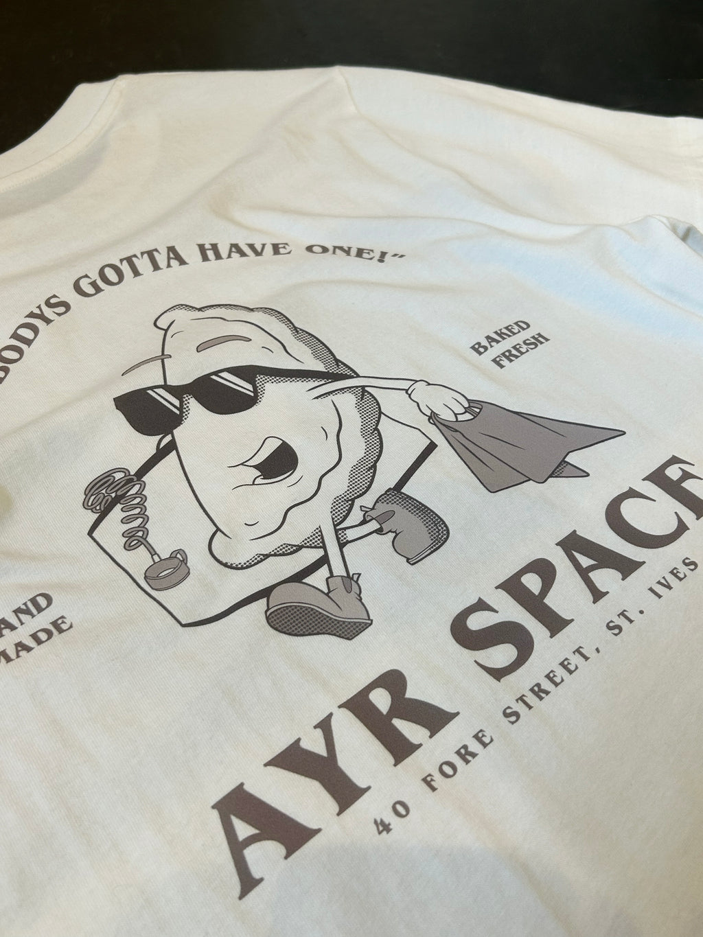 Close-up of the Pasty T back print: faded brown retro cartoon pasty in sunglasses with a paper bag, "Hand made", "Baked fresh" and AYR SPACE, 40 Fore Street, St. Ives