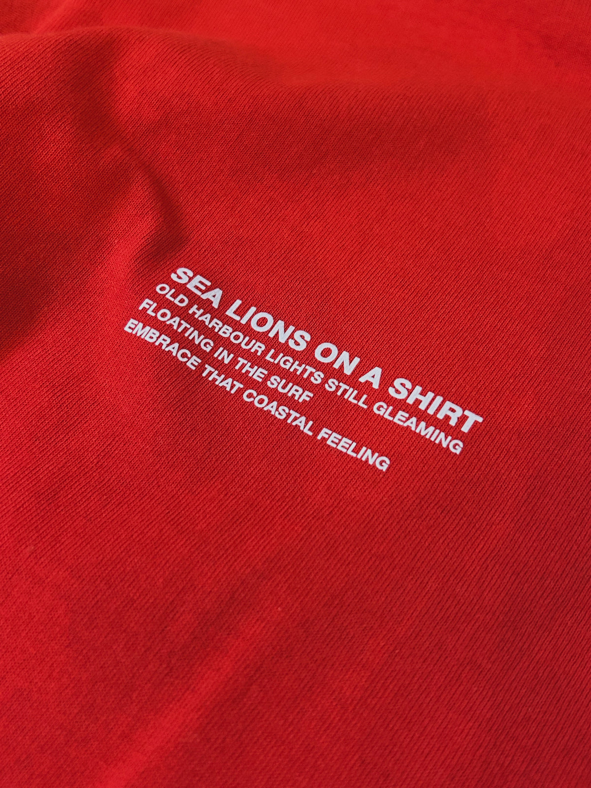 Close-up of the Sea Lions Long Sleeve in Away Red: white chest print reading "Sea Lions on a Shirt - old harbour lights still gleaming, floating in the surf, embrace that coastal feeling"