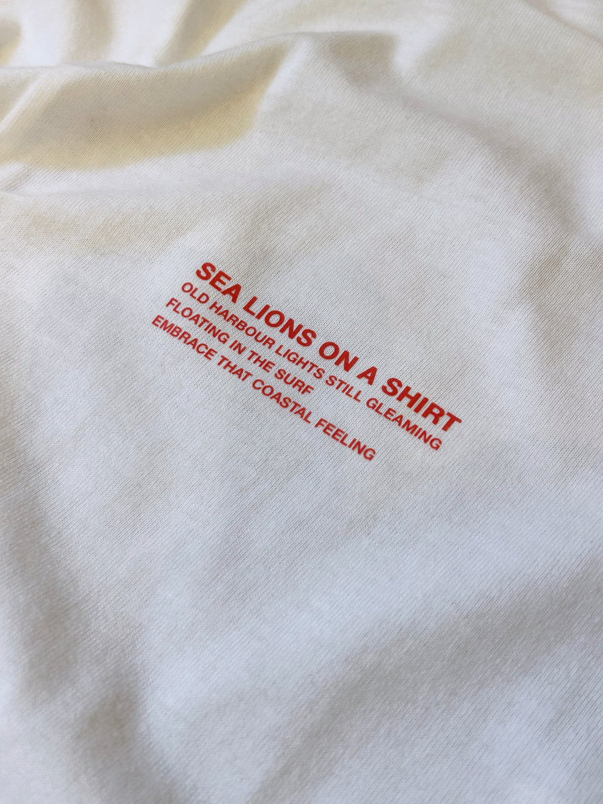 Close-up of the Sea Lions Long Sleeve in Home White: red chest print reading "Sea Lions on a Shirt - old harbour lights still gleaming, floating in the surf, embrace that coastal feeling"