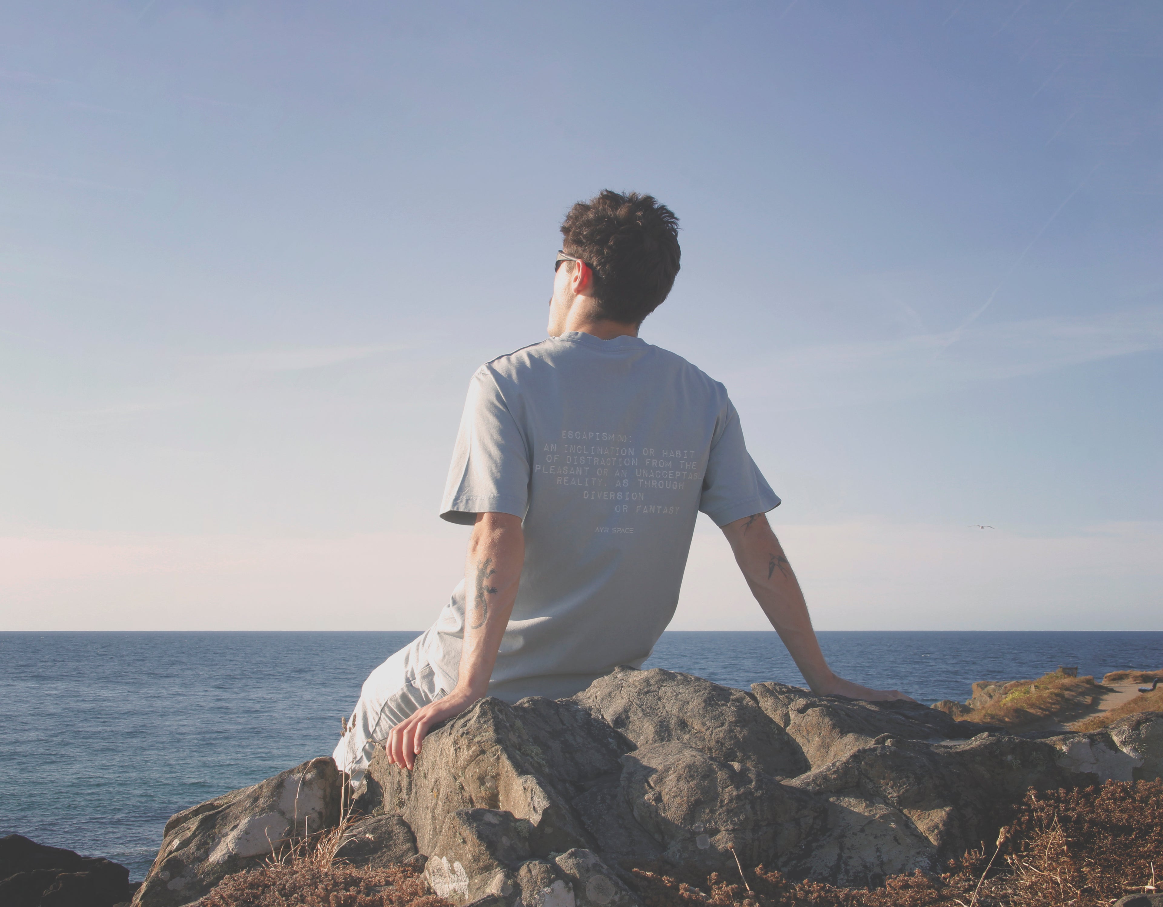 Young man sitting overlooking the coast of St. Ives, Cornwall wearing the Ayr Space Escapism T-Shirt.